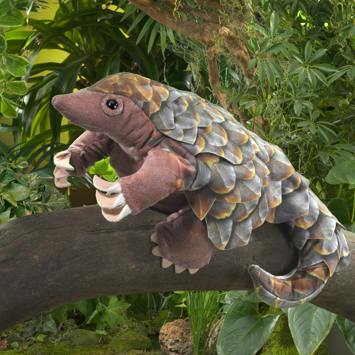 Folkmanis Handpuppe Pangolin mit detailliert gestalteten grauen Schuppen und Krallen, auf einem Ast in tropischer Kulisse
