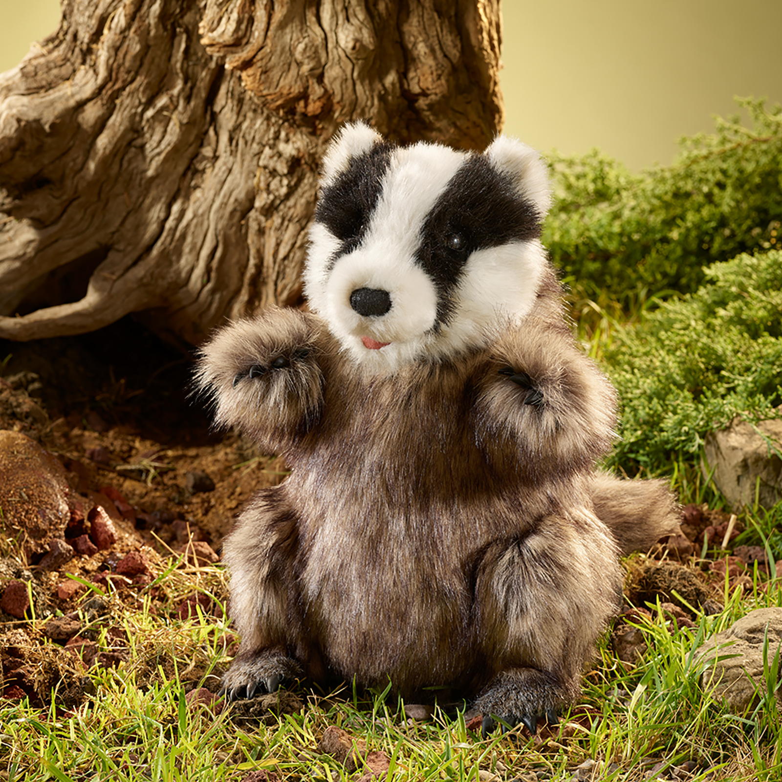 Folkmanis Handpuppe Dachs sitzt vor einem Baumstamm im Gras, mit gestreiftem Kopf und leicht geöffnetem Maul.