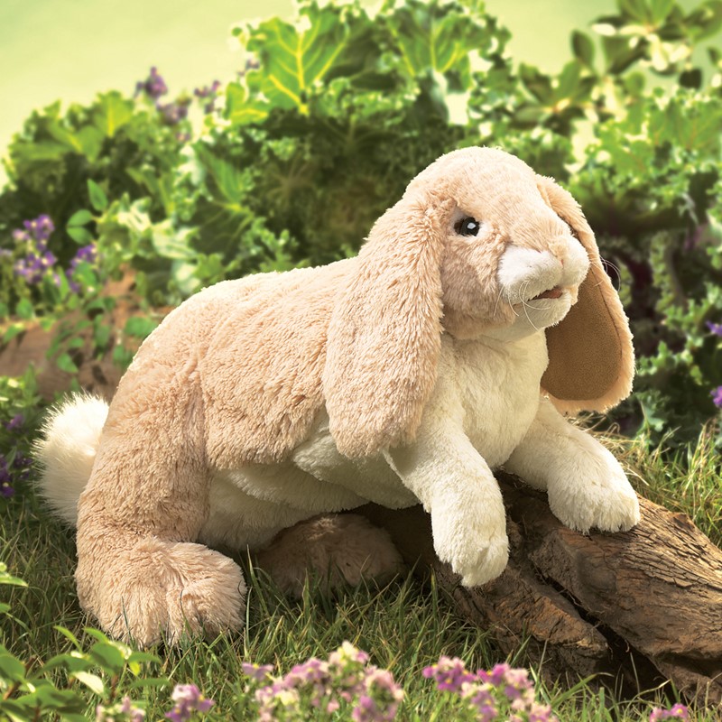 Folkmanis Handpuppe kuscheliges Kaninchen mit hellbeigem Plüschfell und langen Hängeohren, auf einem Baumstamm im Gras liegend