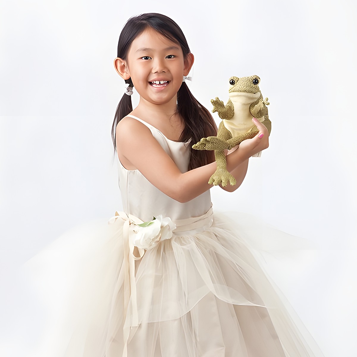 Mädchen im cremefarbenen Tüllkleid hält die Folkmanis Handpuppe Lustiger Frosch in olivgrünem Plüsch in die Kamera.