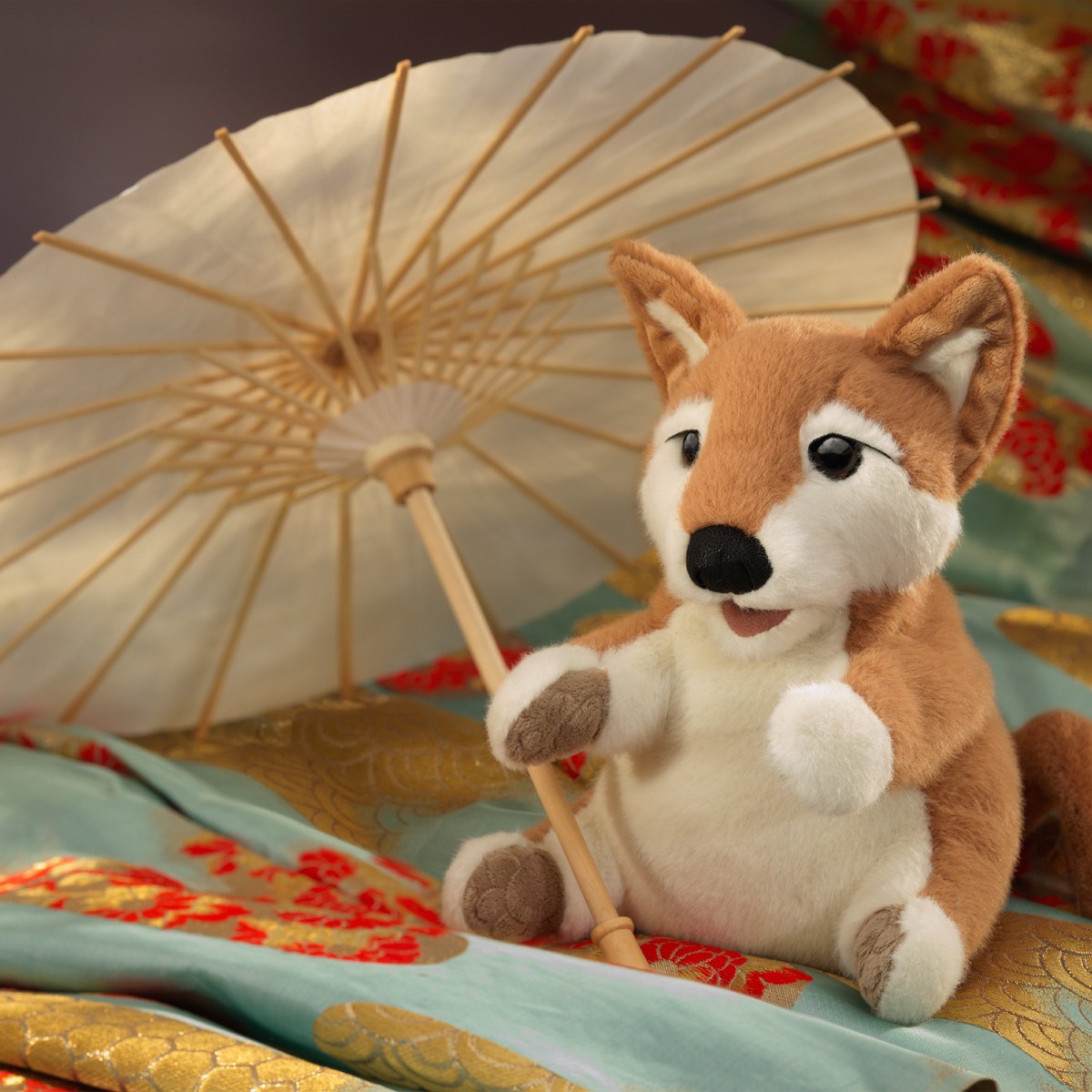 Folkmanis Shiba Inu hand puppet in brown and white, sitting and holding a paper parasol, on embroidered silk fabrics in gold and teal