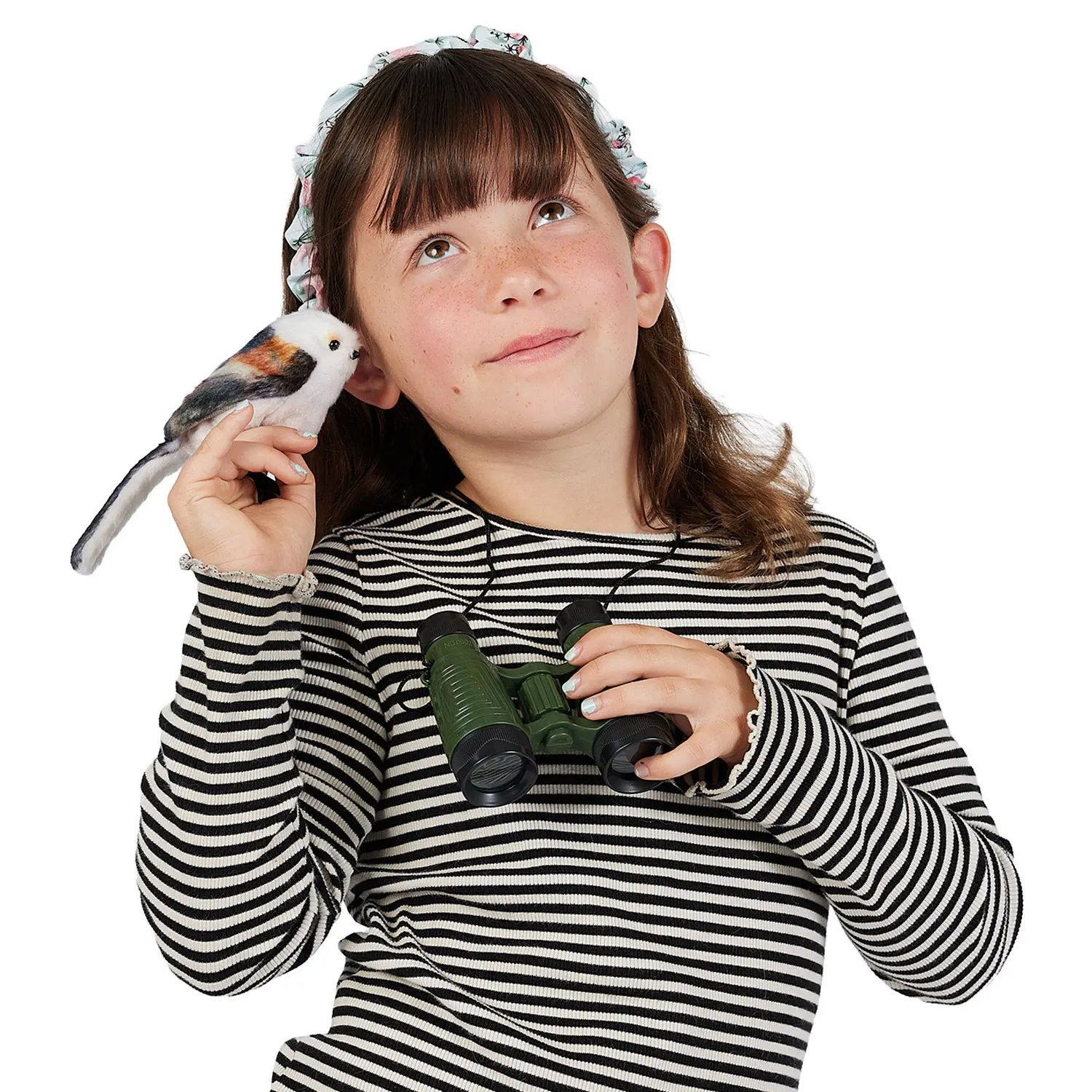 Mädchen hält Folkmanis Fingerpuppe mini Schwanzmeise mit weißem Gefieder und braunen Akzenten neben ihr Ohr, in der anderen Hand ein grünes Fernglas.