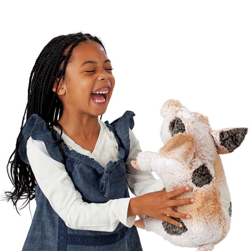 Lachendes Mädchen hält die Folkmanis Handpuppe grunzendes Schweinchen mit beigem Fell und dunklen Flecken in den Händen.