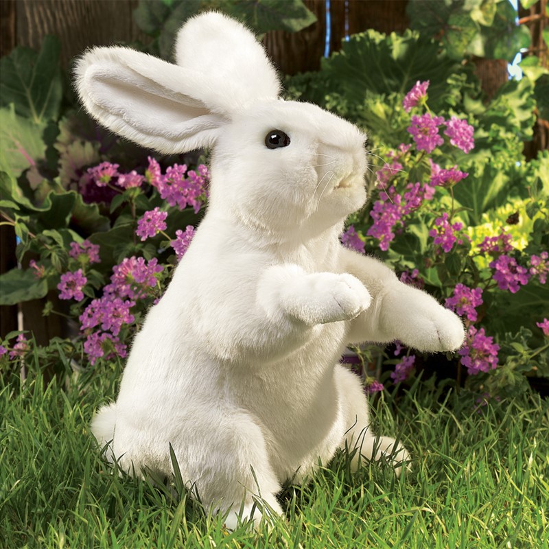 Folkmanis Handpuppe weißes Kaninchen in aufrechter Pose auf Gras, vor lila Blüten, Seitenansicht