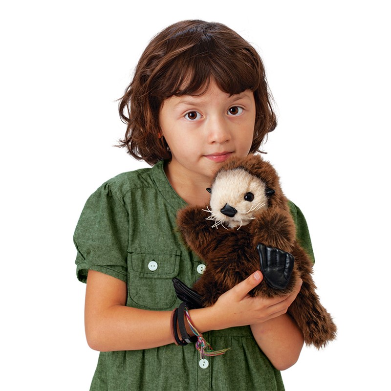 Mädchen hält Folkmanis Handpuppe Seeotter-Baby mit dunkelbraunem Fell und schwarzen Flossen vor weißem Hintergrund.