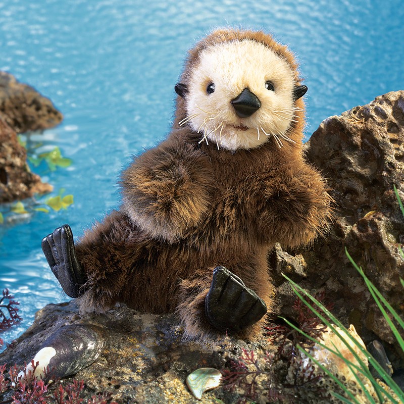 Folkmanis Handpuppe Seeotter-Baby mit dunkelbraunem Fell und cremefarbenem Gesicht, auf Felsen vor blauem Wasser sitzend