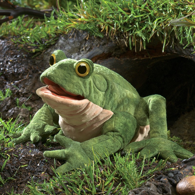 Folkmanis Handpuppe Kröte in sattem Grün mit strukturierter Haut, geöffnetem Maul und gelben Augen, auf feuchtem Boden vor Baumhöhle