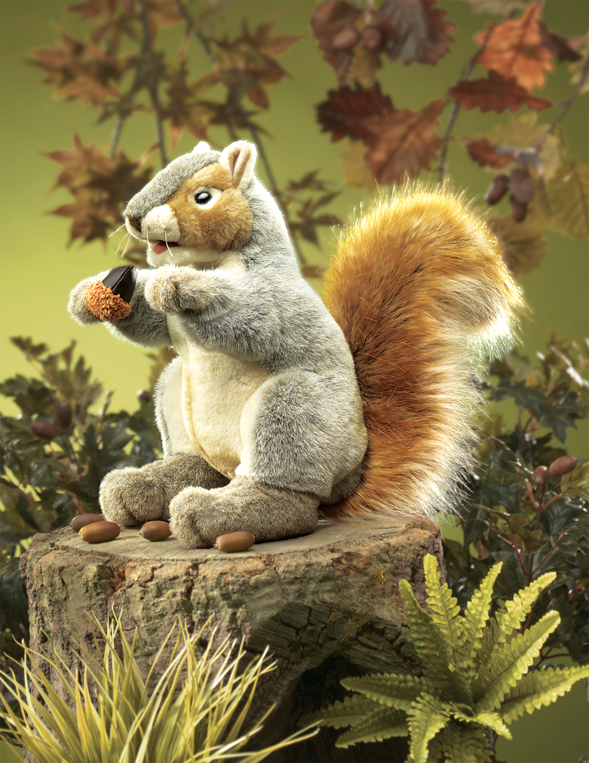Folkmanis Handpuppe graues Eichhörnchen mit orangefarbenem Schwanz hält eine Eichel, sitzt auf Baumstumpf vor Herbstkulisse
