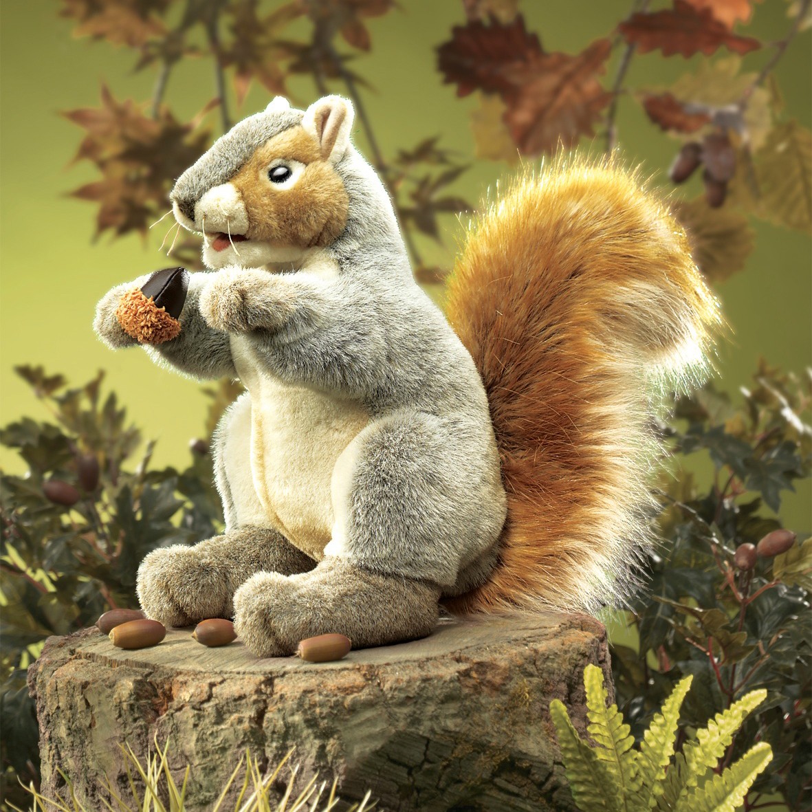 Folkmanis Handpuppe graues Eichhörnchen mit orangefarbenem Schwanz hält eine Eichel, sitzt auf Baumstumpf vor Herbstkulisse