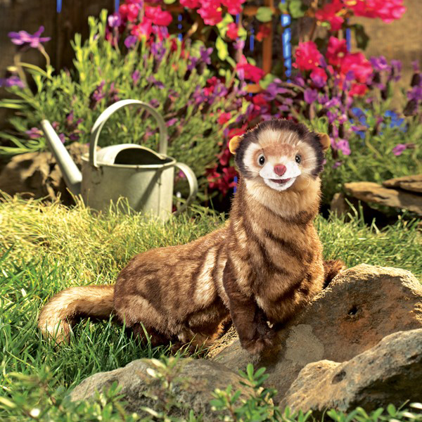 Folkmanis ferret hand puppet with brown and beige fur sitting on a stone, with flowers and a watering can in the background