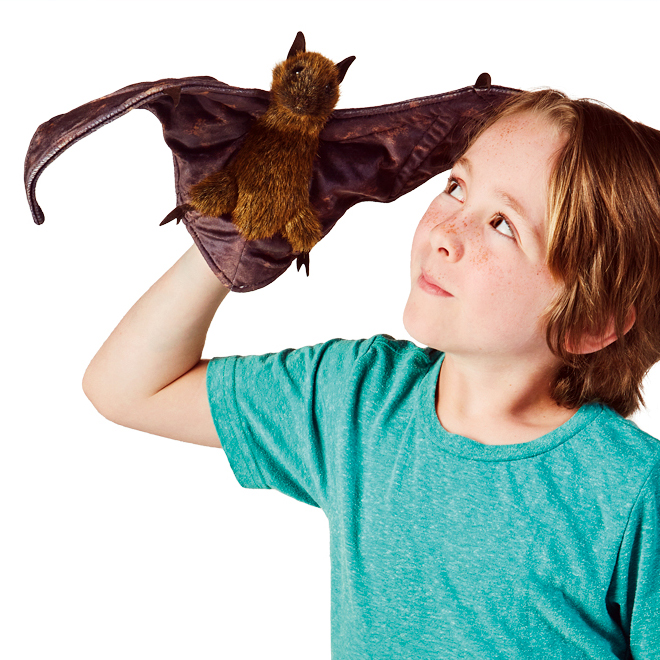 Child holding a Folkmanis fruit bat hand puppet with spread brown leather wings and reddish-brown fur up in the air