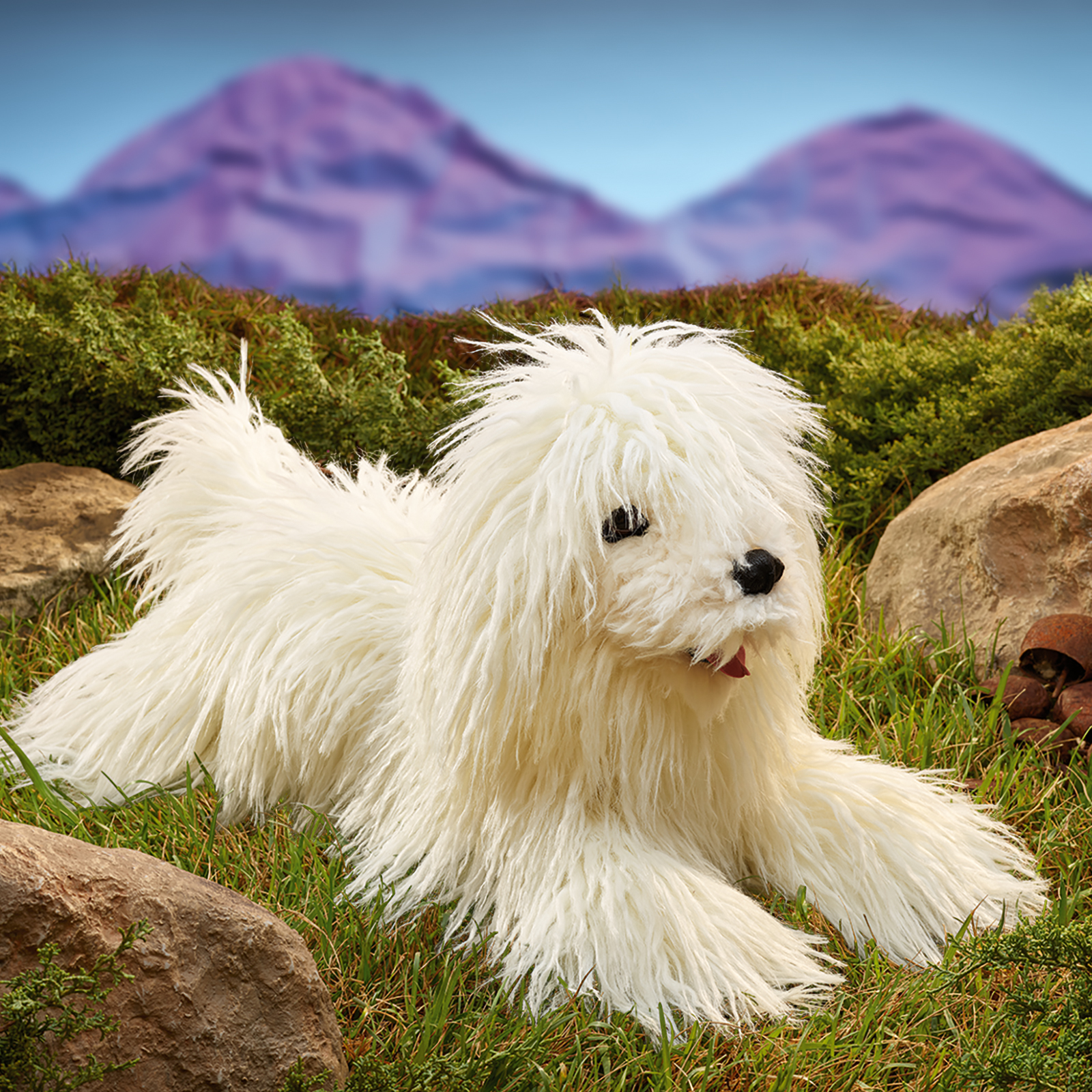 Folkmanis Handpuppe Komondor liegt zwischen Felsen im Gras, das Maul geöffnet.