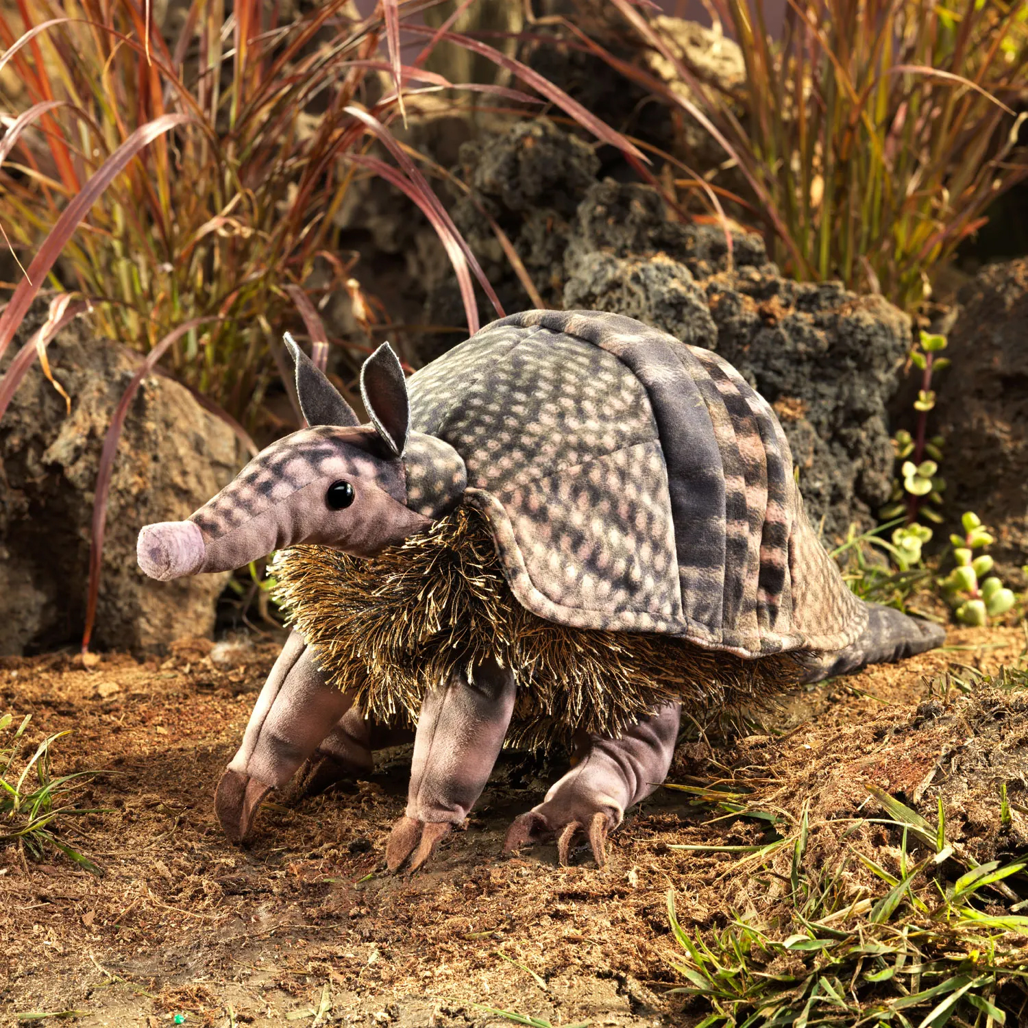 Folkmanis armadillo hand puppet with textured grey-pink shell, pointed snout and golden bristles, side view on sandy ground