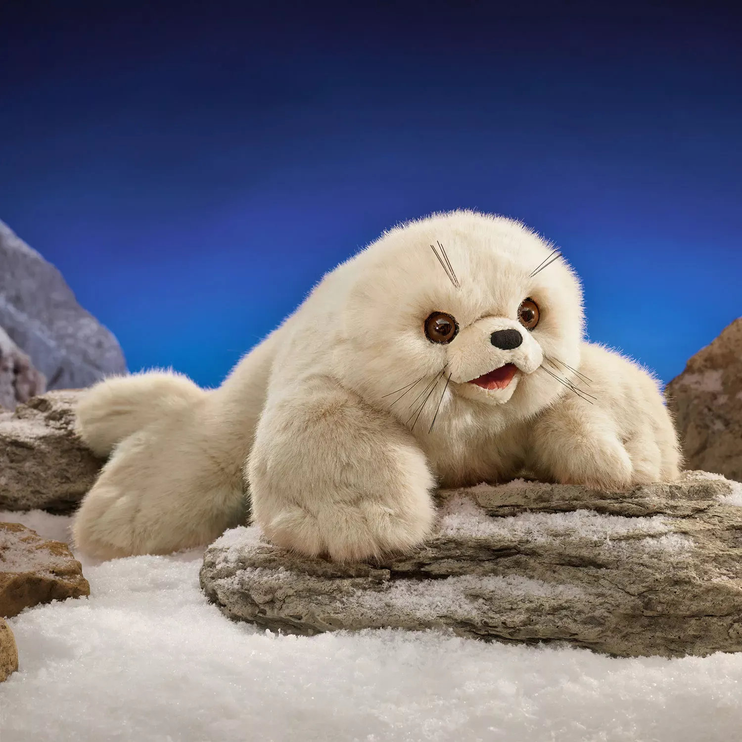 Folkmanis Handpuppe Robbenbaby mit cremefarbenem Plüschfell, geöffnetem Maul und Schnurrhaaren auf schneebedeckten Felsen