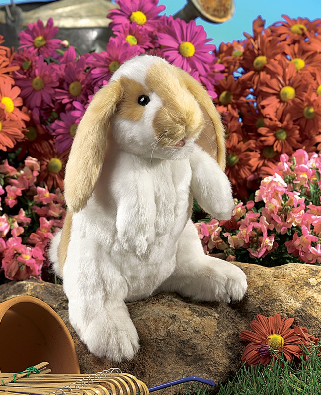 Folkmanis hand puppet standing lop rabbit with white plush fur and beige floppy ears, upright on a stone in front of a floral background