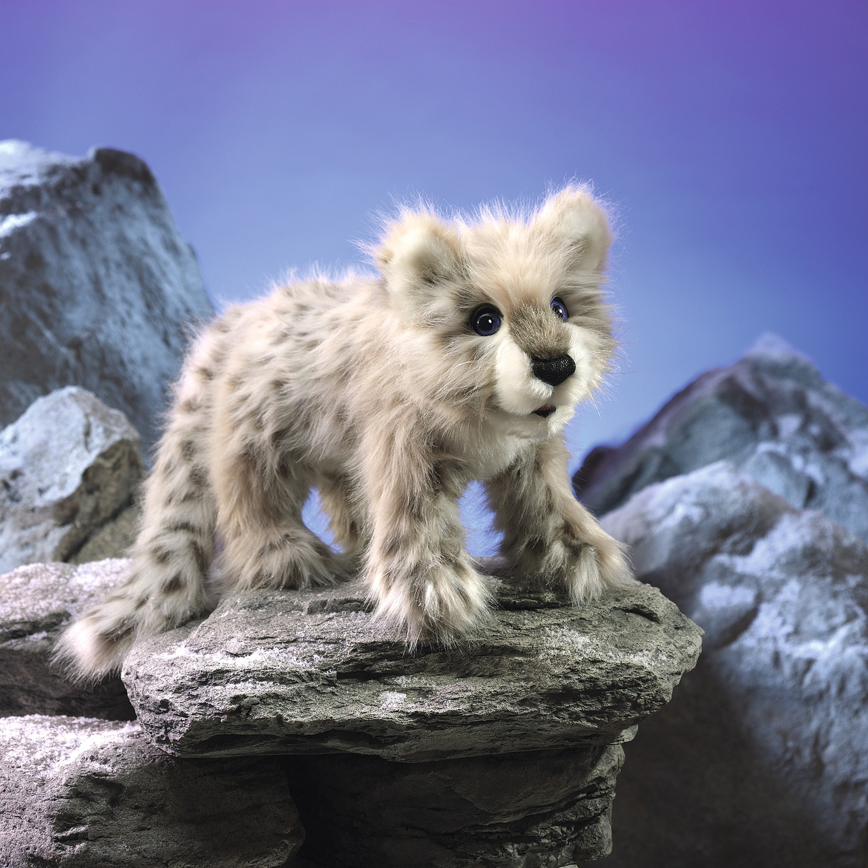 Folkmanis hand puppet baby snow leopard with grey-beige spotted fur, standing on a rock against a blue-violet background